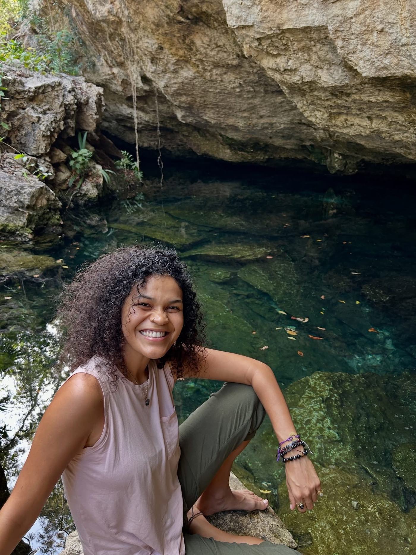 Cyndiii Deusa sitting beside the clear water of a cenote, laughing.