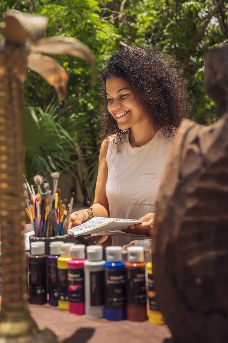 Cyndiii Deusa laughing at a table of paints and brushes, surrounded by green.