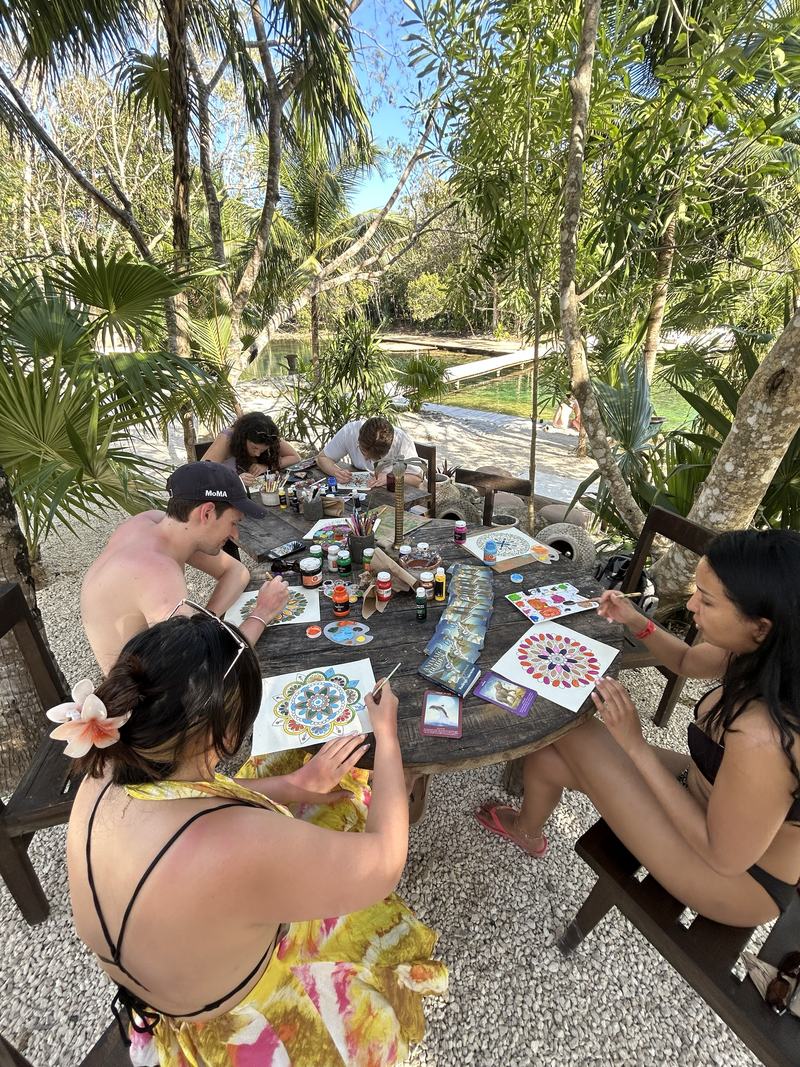 Hands painting mandalas together at a round table beside the lagoon.