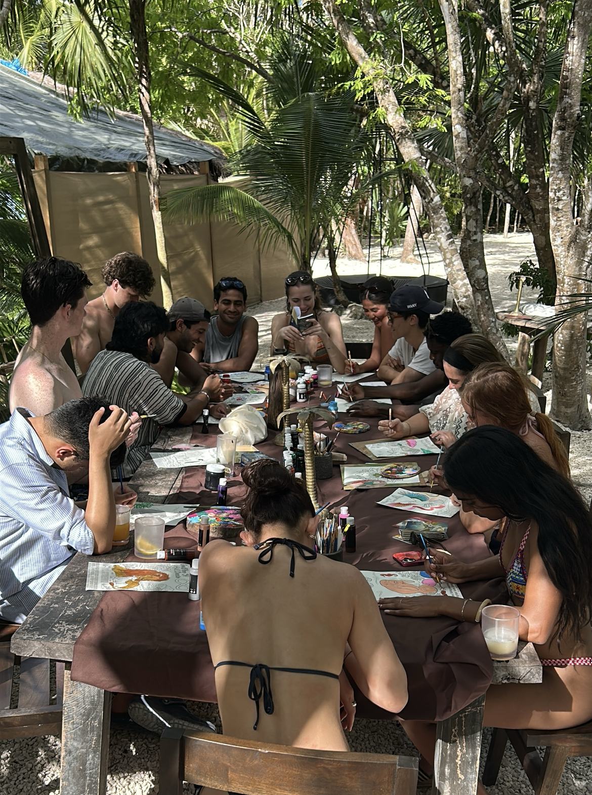 A long table of people gathered together painting by the water, palms overhead.