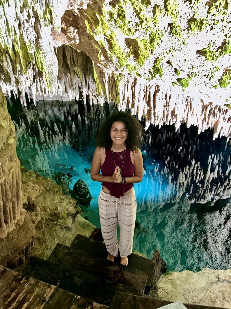 Cyndiii Deusa, hands held in prayer, present beside the cenote's still water.