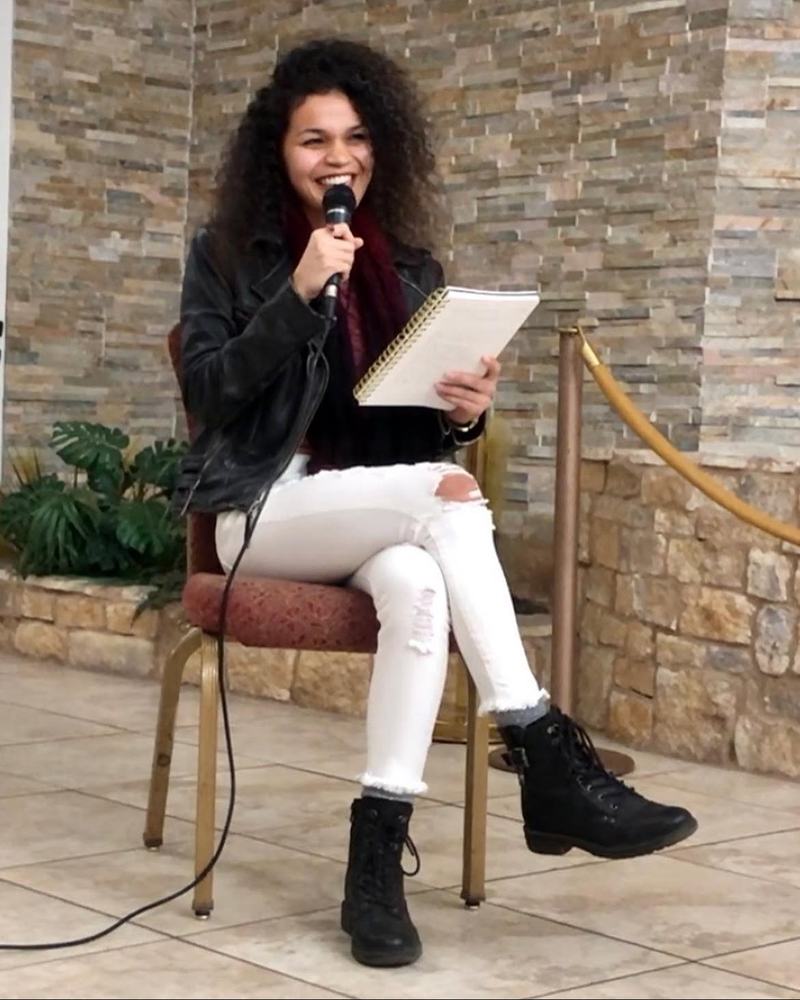 Cyndiii Deusa laughing mid-sentence, microphone in one hand and her poetry book in the other.