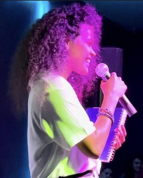 Cyndiii Deusa standing on stage, microphone raised, mid-song under coloured lights.